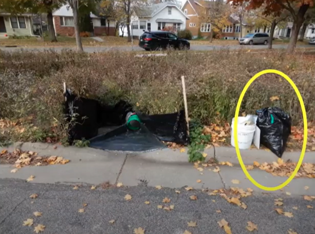 Large, full trash bags sit next to a collection device  made from tarp and tubes installed on a curb cut leading into a dry pond